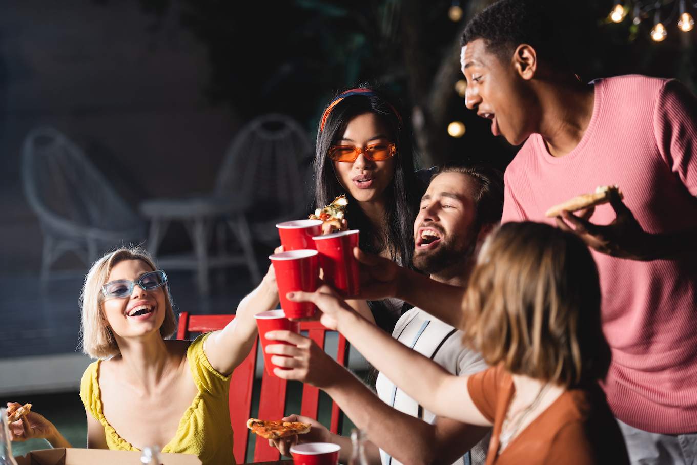 A group of friends toast red plastic cups and eat pizza with chairs and lights in the background.