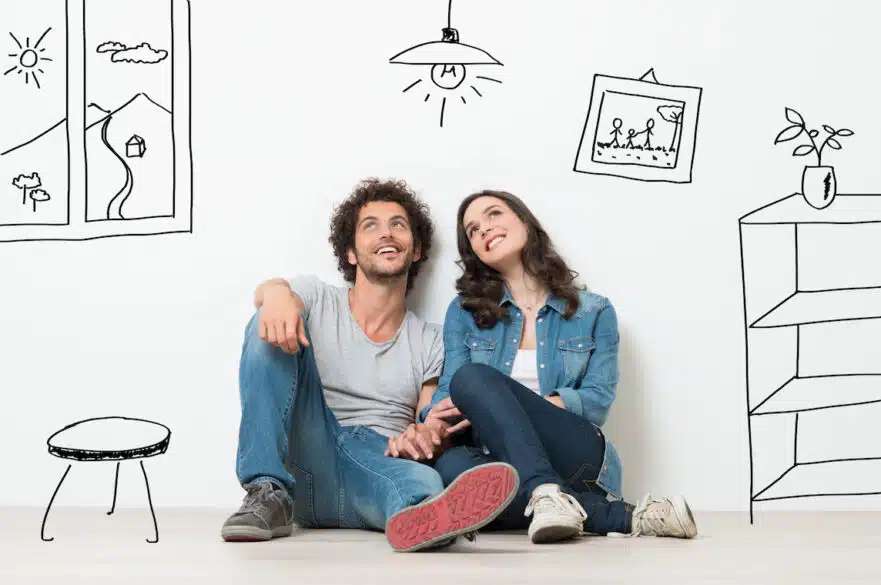 A man and woman imagine the possible furnishings of their new home while sitting against a white wall, clasping hands.