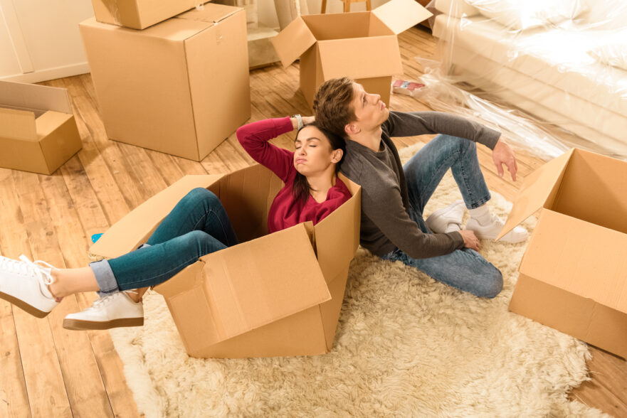 A man leans against a woman sitting in a box. Boxes surround the two, who look tired.