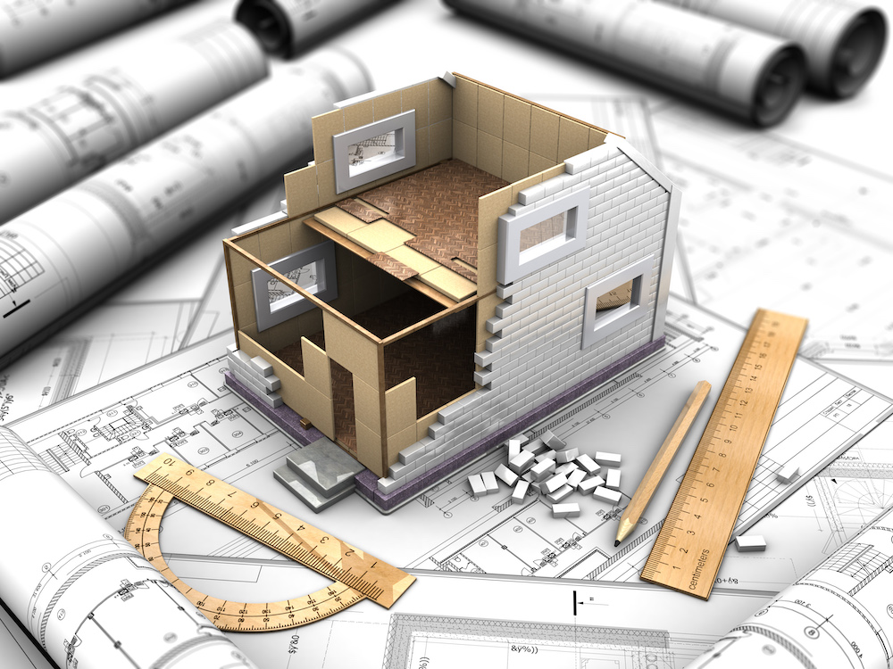 A half-built 3D model of a home sits on top of a two-story house floor plan, surrounded by a pencil, ruler, and protractor.